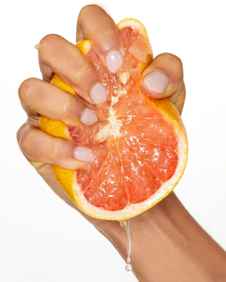Hand squeezing a halved grapefruit with juice dripping from the fruit against a light background.