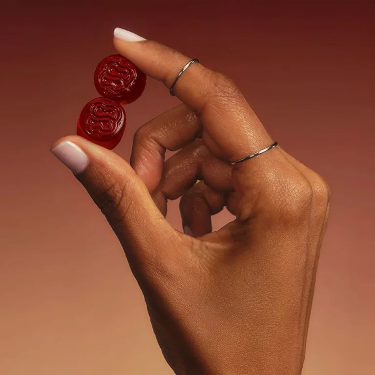 An image of a female hand holding two of Sakara Life's Serene State gummies. The female has silver rings on, and the background of the image is red.