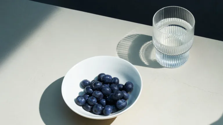 Small bowl of blueberries beside a glass of water and a cup of black coffee on a white table.