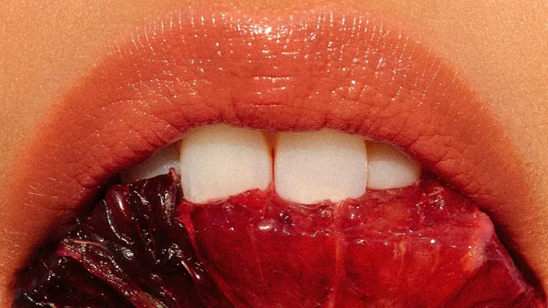 Close-up of woman's mouth with glossy lips taking a bite out of a blood orange.