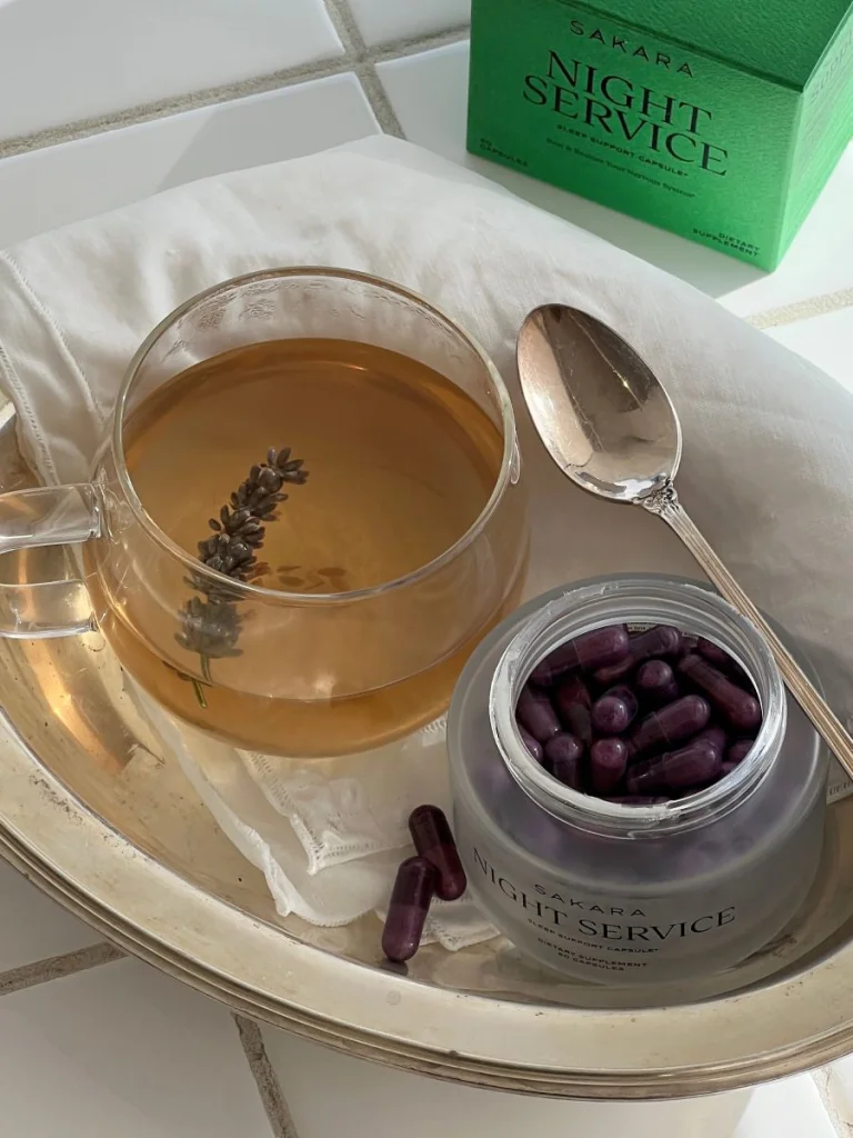 A clear glass mug with tea and a lavender stem next to Sakara Night Service capsules in a frosted jar, as well as the green Night Service box packaging.