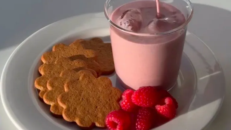 A glass of pink-colored strawberry milk on a plate with cookies and raspberries.