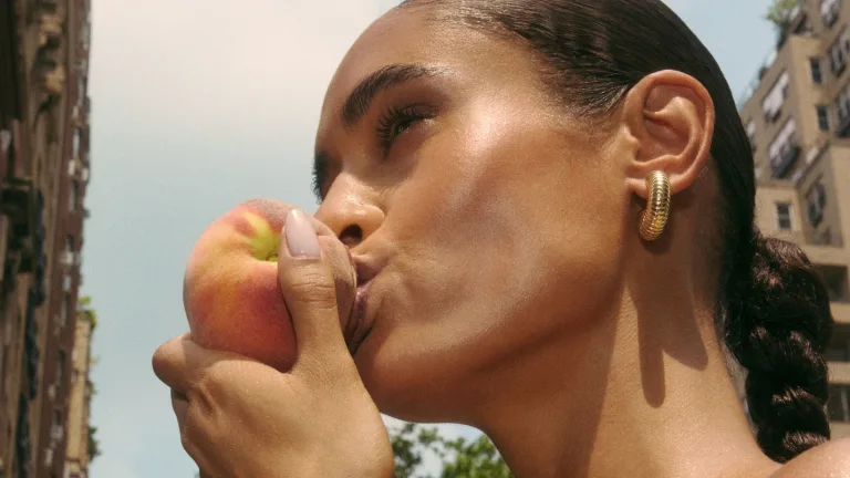 A close-up image of a woman taking a bite out of a peach, with a background of NYC.