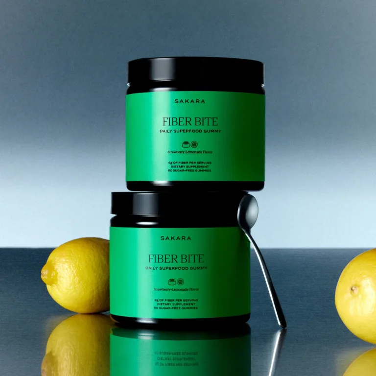 Two stacked Sakara Fiber Bite daily superfood gummy jars in black packaging with bright green labels, styled on a reflective surface with whole yellow lemons and a metal spoon against a soft blue gradient background.