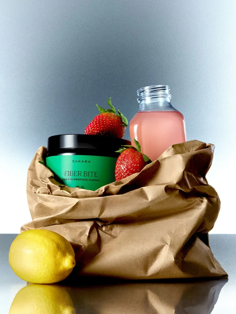 Sakara Fiber Bite, strawberries, lemons, and a glass bottle with pink liquid inside a brown paper bag on a blue background