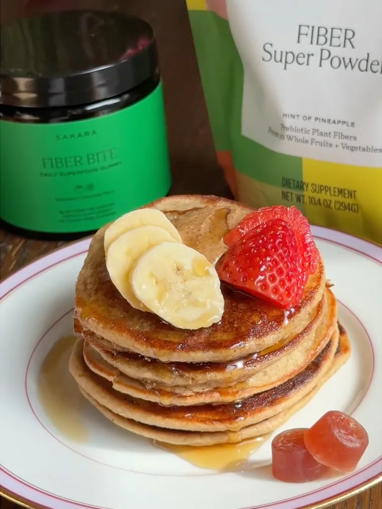 Three-step collage showing Sakara Fiber Super Powder and Fiber Bite used to make pancakes. Left: a hand pours tan fiber powder from a small metal cup into a clear blender filled with oats and liquid, with a green Sakara Fiber Bite jar and yellow-and-white Fiber Super Powder pouch visible in the background. Center: pancake batter cooking in a black skillet, with a spatula lifting a golden-brown pancake. Right: a stack of fluffy pancakes topped with sliced banana, strawberry, and syrup on a white plate, with the green Fiber Bite jar and Fiber Super Powder pouch displayed behind on a wooden surface.