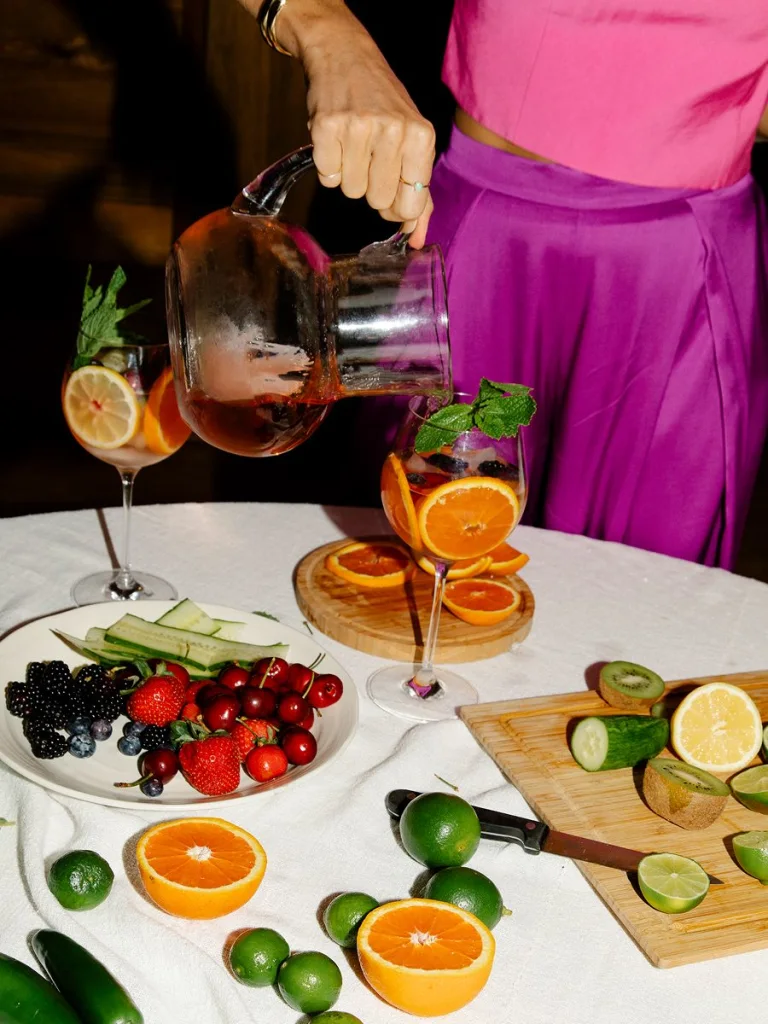Close-up of a woman in a bright pink satin top and skirt pouring a citrus spritz from a clear glass pitcher into a stemmed wine glass garnished with orange slices and fresh mint. The table is styled with sliced oranges, limes, cucumber, kiwi, and a plate of strawberries, cherries, blackberries, and blueberries on a white tablecloth.