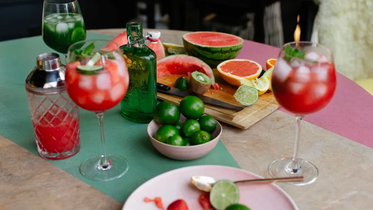 Close-up of a styled cocktail table featuring two red watermelon cocktails in stemmed glasses with ice and mint, surrounded by fresh ingredients including whole limes, sliced citrus, kiwi, and a halved watermelon on a wooden cutting board, with a green glass bottle and cocktail shaker nearby.