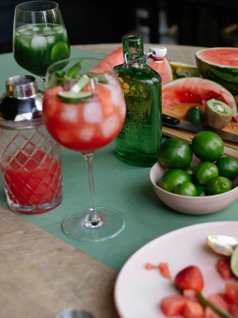Close-up of a styled cocktail table featuring two red watermelon cocktails in stemmed glasses with ice and mint, surrounded by fresh ingredients including whole limes, sliced citrus, kiwi, and a halved watermelon on a wooden cutting board, with a green glass bottle and cocktail shaker nearby.