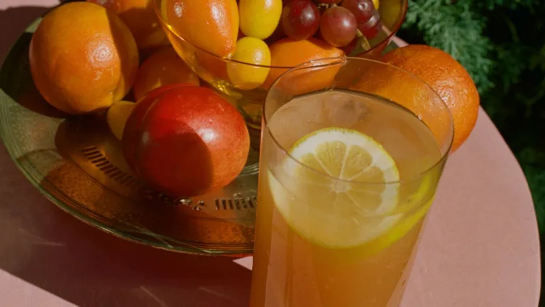Close-up of a citrus drink in a clear glass with a lemon slice, set on a pink table beside a glass bowl filled with fresh fruit including oranges, grapes, kumquats, and a nectarine, with warm sunlight casting rich shadows.