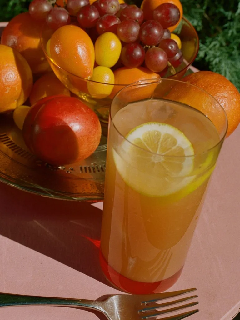 Close-up of a citrus drink in a clear glass with a lemon slice, set on a pink table beside a glass bowl filled with fresh fruit including oranges, grapes, kumquats, and a nectarine, with warm sunlight casting rich shadows.