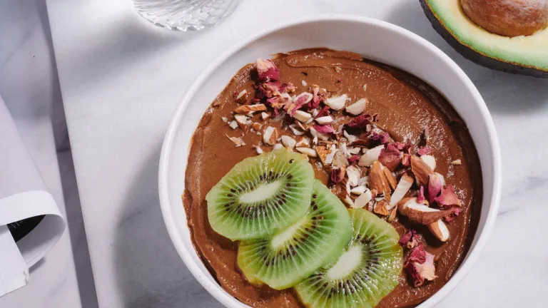 Overhead view of a creamy chocolate avocado pudding in a white bowl, topped with sliced kiwi, chopped almonds, coconut flakes, and dried rose petals, with a halved avocado, a small bowl of cocoa powder, and a glass of water arranged on a light marble surface.