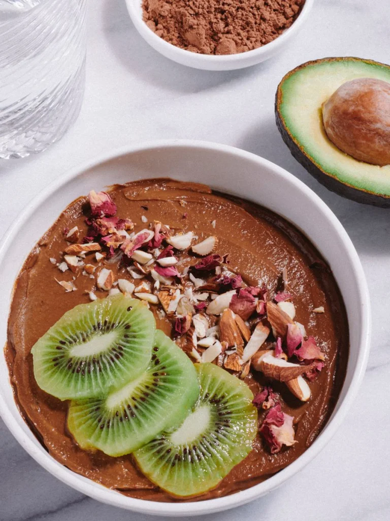 Overhead view of a creamy chocolate avocado pudding in a white bowl, topped with sliced kiwi, chopped almonds, coconut flakes, and dried rose petals, with a halved avocado, a small bowl of cocoa powder, and a glass of water arranged on a light marble surface.
