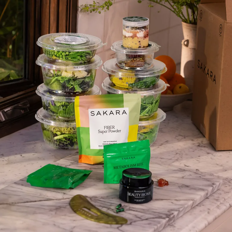Plastic containers of Sakara salads stacked on counter with Fiber Powder, Metabolism Bite and Beauty Biome in Front 