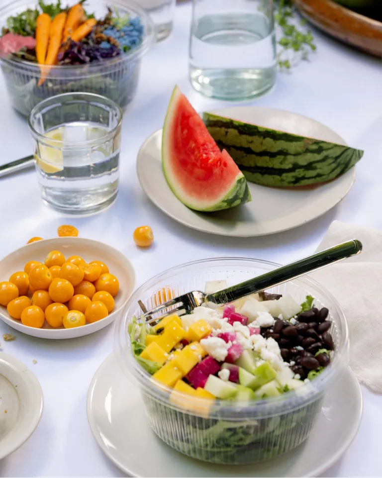 Sakara salad next to slices of watermelon on a table with white tablecloth and water cups